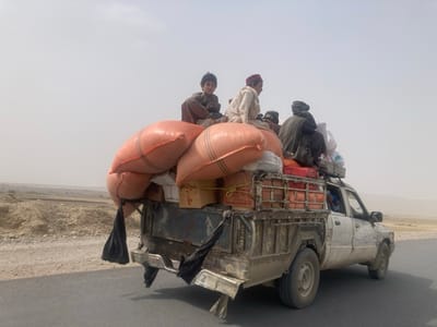 A heavily laden, dirty white pickup truck travels on a paved road under a hazy sky. On top of the cab and truck bed are numerous large, orange sacks, boxes, and caged containers. Several people, including children, sit atop the precarious load. The truck’s tailgate is obscured by dark material, possibly cloth. A barren landscape stretches into the distance on either side of the road. Dust and grime coat the vehicle.