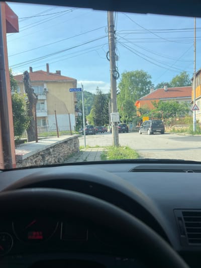 The image, taken from inside a vehicle, shows a street scene in Breznik, Bulgaria. A utility pole with numerous wires dominates the left side of the frame. Buildings line the street, with houses featuring orange tile roofs. Parked cars are visible along the right side of the road. A street sign reading "ул. Kpan Cesse Str. Krom Serev Str." is present on a building. A portion of the car's dashboard and steering wheel are visible in the foreground.