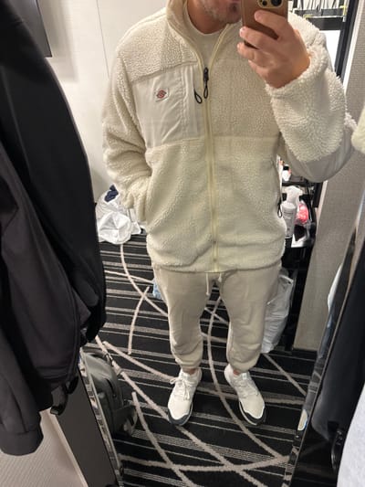 The image shows a man standing in a hotel room in Chiyoda City, Tokyo, Japan. The man is wearing a white fleece jacket with a zipper, and cream-colored pants. He is holding a smartphone in his right hand. He is wearing white sneakers on his feet. He is standing in front of a mirror, and there are other items, including a black jacket and a bag, near him. There is a black and white patterned carpet on the floor. The room appears to be a hotel room with a mirror, luggage rack, and a closet door. The man is standing on a patterned carpet and holding his phone. He is wearing white sneakers and a cream colored outfit. The location is a hotel room in Tokyo, Japan. The location's coordinates are latitude 35.675, longitude 139.741.