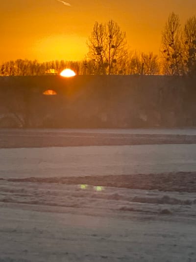 A blurred image is divided into three distinct sections. The left depicts a sunset with golden light filtering through bare trees. The middle section is a dark, ambiguous, vertical area with a single, small, orange reflection. The right-most part appears to be a frosted or textured surface with faint vertical patterns. Two small, bright spots are visible near the center of this section. Overall, the image has an indistinct and possibly photographic quality.