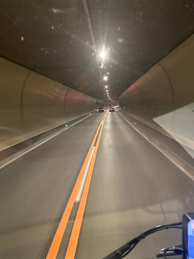 The image captures the interior of a tunnel, likely a road tunnel, with a perspective looking forward. The tunnel is characterized by its curved, beige-colored walls and a dark ceiling from which lights are visible, creating streaks of light due to motion blur. The road surface is asphalt, marked with double orange lines in the center, indicating lanes of traffic. Several cars are visible in the distance, moving in the same direction. The foreground shows a portion of a dark-colored handlebar, suggesting the image was taken from a vehicle, possibly a motorcycle or a scooter. The overall impression is one of transit through an enclosed, artificial environment. The specific location in Lindau (Bodensee), Germany, is not determinable from the visual elements alone.