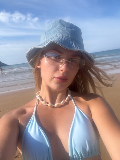 A young woman, wearing a light blue bikini top, a white shell necklace, and a denim bucket hat with a Nike logo, takes a selfie on a beach. She is also wearing rimless sunglasses with a slight tint. The background shows a sandy beach, calm ocean water, and a bright blue sky with scattered white clouds. Two people are visible in the shallow water in the distance. The image is taken outdoors during the daytime. The location is not Hoa Xuan, Vietnam, but rather a generic beach setting.