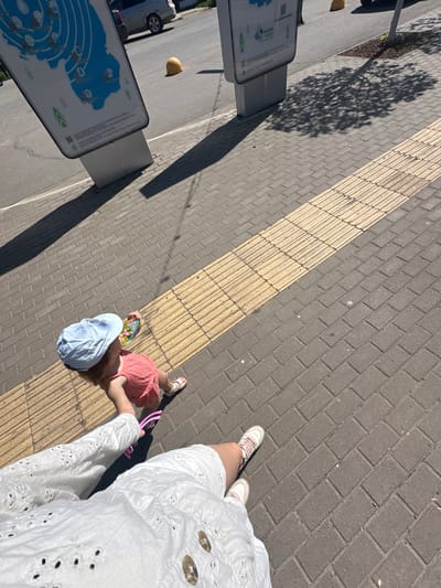 A high-angle shot shows two children lying on a sidewalk. One child wears a light blue hat and holds a book. The second child is next to the first wearing a red dress and sandals. A woman wearing a white dress stands over the children, holding a pink object. A sidewalk with textured yellow stripes lies beneath the children. A tree’s shadow falls on the sidewalk. Two signs are on the left side of the image. The location is in the city of Uva, Russia.