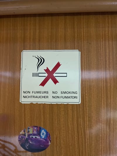 The image shows a "No Smoking" sign with corresponding translations in French, German, and Italian. The sign is rectangular, white with a black border. It has a cigarette symbol with a red "X" over it, along with wavy lines representing smoke. Below the symbol are the words "NON FUMEURS" "NICHTRAUCHER" "NON FUMATORI". To the right of the sign is a small round sticker featuring a purple car from the Pixar movie "Cars". The background is a light brown surface with a wood grain pattern. There is insufficient information to determine if the image was taken in the city of Gornje Mrke, Montenegro. The image is a close-up shot of these items, obscuring the wider environment.