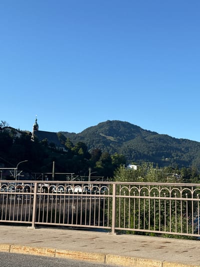 This is a landscape photograph taken from a bridge overlooking a town nestled in a valley with a mountain range in the background. The sky is clear and blue. In the mid-ground, there is a town with buildings, including a church with a distinctive spire. The town is situated on a hillside covered in dense green trees. The mountain range behind the town is also covered in dark green coniferous trees. The foreground of the image features a concrete walkway and a decorative metal railing. Beneath the railing, there are train tracks and some vehicles visible. The overall impression is that of a picturesque mountain town on a sunny day. The image appears to be taken in Berchtesgaden, Germany, given the presence of a church spire and the mountainous terrain characteristic of the region.