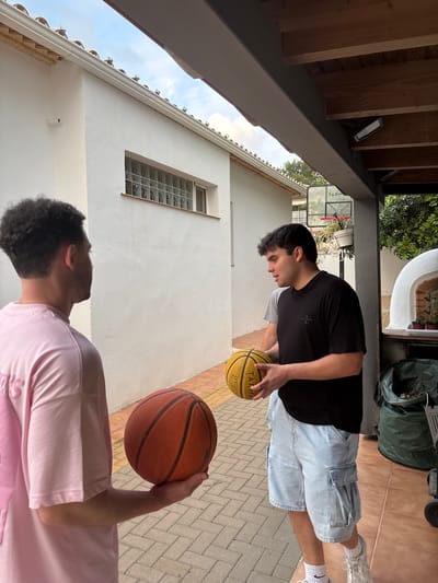The image displays an outdoor scene featuring two young men engaged in a basketball activity on a paved patio. The setting is bright and appears to be daytime, with a partly cloudy sky visible.
In the foreground, a man with dark, curly hair, wearing a pink t-shirt, stands with his back partially to the camera. He holds an orange basketball in his right hand and is looking towards the second man. The second man, positioned in the mid-ground, has straight dark hair and is wearing a black t-shirt with a small, light-colored emblem and light blue denim shorts. He is focused on a yellow basketball, which he holds with both hands. His white socks display a black Nike swoosh.
The background shows a white-walled house with a distinctive narrow, rectangular window composed of glass blocks. Above, a wooden beamed ceiling extends over a portion of the patio, equipped with two black floodlights. A basketball hoop with a clear backboard is visible behind the man in the black shirt. The backboard has "TARMAK" printed on it. Lush green foliage and bushes are also seen behind the house and hoop. Near the back, a dark green bag and a wheeled object are present. The yellow basketball held by the second man bears the text "TARMAK BT100 SOFT." The scene suggests a casual practice or game in a residential outdoor area, consistent with a patio in Montgó, Spain.