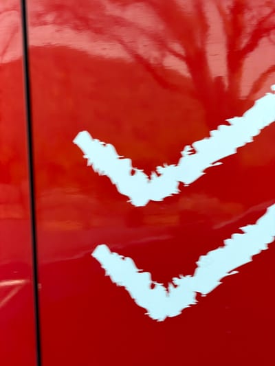 A close-up reveals two stylized, jagged white arrows pointing right against a glossy red background. The background is somewhat reflective, showing blurry reflections. The arrows appear slightly distressed, with uneven edges that give them a rough, almost torn appearance. The bottom edge of the frame is bordered by a black line.