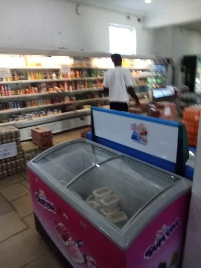 The image shows the interior of a store located at 5.878, 5.699 in Sapele, Nigeria. The scene features a large freezer with a glass lid and a pink and white exterior. It contains white containers. Behind the freezer, a man in a white shirt walks past shelves stocked with packaged goods. There is another freezer in the background. The walls and floor are made of light-colored tiles. A sign on the back of the freezer advertises a product. The space is well-lit and appears to be a store with a variety of items for sale.
