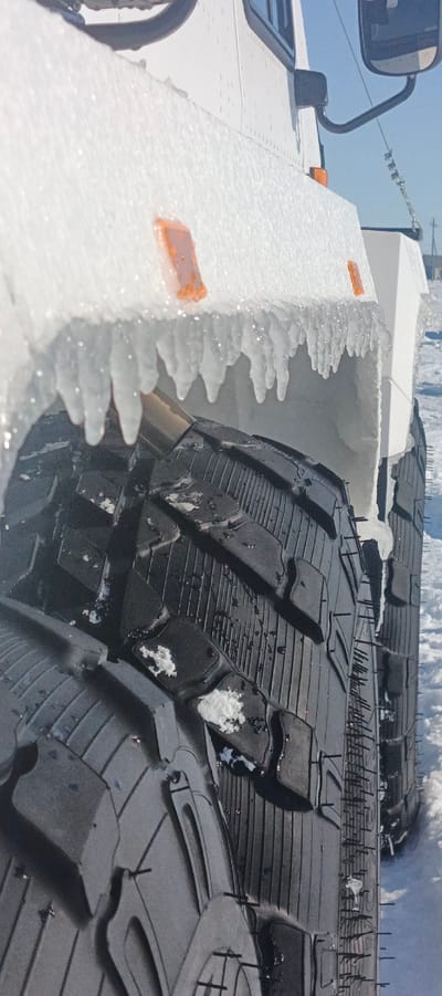 A white heavy-duty vehicle is observed in a snowy outdoor environment in Nelmin-Nos, Russia. The vehicle's lower side body panel and wheel well are significantly encrusted with ice, with numerous clear to opaque icicles, approximately 5-15 cm in length, hanging from the underside of the fender lip. Two amber rectangular marker lights are visible on the ice-covered side panel. Two large, dark-colored, studded tires are partially visible, positioned on a snow-covered surface. The tires feature an aggressive block tread pattern, and metallic studs are clearly visible protruding from the tread blocks. Patches of snow and ice remain within the tire treads. The background consists of a bright, clear sky above a flat, snow-covered landscape with indistinct distant structures, indicating freezing temperatures and winter conditions. No individuals are present.