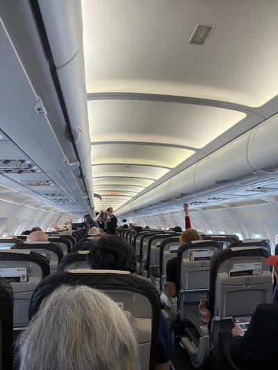The image depicts the interior of an airplane cabin, seen from a passenger's perspective looking down the aisle. The original image is rotated 90 degrees counter-clockwise; when oriented correctly, the aisle runs horizontally from foreground to background, with rows of seats on either side.
**Subjects:** Numerous passengers are visible, mostly from behind, seated in rows along the aisle. One individual with grey hair is prominent in the immediate foreground. Further down, a person with dark hair is visible, and others are seen in various seats. A person in a red long-sleeved top is captured reaching up into an overhead compartment. Another passenger is seen holding a tablet.
**Objects:** The cabin features rows of grey and black upholstered airplane seats. Overhead compartments line both sides of the cabin. Bright, diffused strip lighting illuminates the ceiling and upper walls. Passenger service units with individual lights and air vents are visible above the seats. Magazines, some bearing the "SWISS" logo, are tucked into seatback pockets. A small, colorful object, possibly a toy or personal item, is resting on a seat.
**Activity or event taking place:** Passengers are either boarding, settling into their seats, or retrieving items from overhead bins, indicated by the person reaching up. Others appear to be relaxing or engaged with personal devices. The scene suggests a typical moment during air travel, either before takeoff or after landing, or during the flight.
**Notable details:** The cabin is predominantly white and grey, giving it a clean and modern appearance. The bright artificial lighting provides a clear view of the interior. The external location context provided is Kloten, Switzerland, suggesting a Swiss airport.
**Visible text:** The word "SWISS" is clearly visible on magazines in the seatback pockets, indicating Swiss International Air Lines. An "EXIT" sign is visible further down the aisle on the side wall. Additionally, "A B C" markings are seen on the overhead passenger service units, likely indicating seat assignments, and the number "29" is visible on the latch of an overhead compartment.
