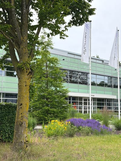The image depicts the exterior of the Neways facility located in Son en Breugel, Netherlands. The building features a modern design with extensive green-tinted glass windows and white framing. In front of the building, there are two white flags with a blue logo and text, possibly company branding.

In the foreground, a prominent, weathered tree with mossy bark stands to the left. Behind it, a meticulously landscaped garden area is visible, characterized by a vibrant bed of purple flowers and yellow flowers, interspersed with green foliage and tall ornamental grasses. A hedge runs along the left side of the image, bordering the garden and the building. The overall environment suggests a well-maintained corporate or industrial setting in a suburban or semi-rural area. The sky is overcast, indicating a cloudy day. No individuals are clearly visible in the frame.