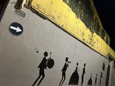 The image depicts a night scene, illuminated by artificial light, showcasing a mural and various urban details on a wall. The wall is predominantly light grey on its upper section, featuring a series of black human silhouettes marching in profile from left to right. These figures appear to be part of a musical procession or parade.
The silhouettes include individuals holding musical instruments such as a trumpet and a large drum with a drumstick. Another figure carries what seems to be a bouquet of flowers or branches. Each figure has distinct headwear or hairstyles. A small, reversed signature or tag, "Est jill," is visible below the last silhouette on the right.
The lower portion of the wall, and extending into the foreground, is painted with diagonal yellow and black stripes. This section shows significant wear and tear, with much of the paint peeling and cracked, revealing a darker surface beneath. Electrical cables run along the top edge of this yellow and black section.
On the right side of the grey wall, a circular dark blue sign with a bold white arrow pointing left is visible. Below it, a rectangular white street sign reads "Carrer Alcalde Juan Bautista Company" in black text. Below this, in smaller text, it states "Ajuntament de Benifassot." On the far left, a portion of a white building with a window and a reversed sign displaying "JOLAN" can be seen. The overall lighting suggests the photograph was taken at night.