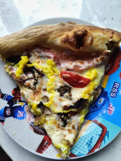 A close-up, overhead shot captures a single slice of pizza on a white plate with a blue, hockey-themed design. The pizza has a thick, golden-brown crust with some charred spots. The toppings include melted white cheese, sliced mushrooms, what appears to be ham or prosciutto, and bright yellow sauce, possibly hollandaise or a cheese sauce, with some pieces of tomato. The plate's design features hockey players, pucks, and a net. The background is a textured white surface, possibly a table or countertop, with natural light filtering in from the left. There is no indication of the city of Petrovsk, Russia, in the image.
