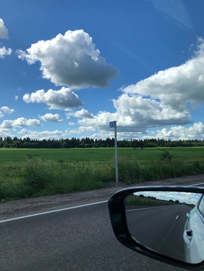 The image depicts a rural roadside scene with a vast green field stretching towards a dense forest line under a blue sky filled with fluffy white clouds. A road runs along the bottom of the frame, with the reflection of the sky and landscape visible in the side mirror of a car. A blue sign with the number "23" is planted on a pole next to the road. The car appears to be moving, as indicated by the reflection in the mirror. The environment suggests a summer day in a natural setting, possibly in the city of Krasnyi Bor, Russia, given the prevalence of forests and open fields. There are no people visible in the image.