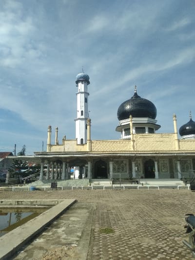The image displays the exterior of Mesjid Punteut in Mesjid Punteut, Indonesia, under a bright, partly cloudy blue sky. The mosque structure, constructed from yellowish-beige stone or concrete, features intricate geometric patterns on its upper facade. Two prominent black, ribbed domes, each topped with a crescent moon and star finial, are visible; one is larger and centrally located, while another smaller dome is partially visible to its right. To the left of the main dome stands a tall, slender white minaret, distinguished by small rectangular windows and topped with a blue, onion-shaped dome.

The mosque's ground level presents a portico supported by multiple white columns, leading to arched entrances. A series of white barriers or railings delineate the immediate area in front of the mosque's steps. In the foreground, a large, open courtyard paved with interlocking hexagonal pavers extends towards the mosque. To the left, a rectangular concrete basin containing still water reflects the sky and distant trees, with a concrete walkway or divider spanning its width. A small patch of green vegetation is visible within the paved area. On the far right edge of the courtyard, the front wheel and part of the engine of a dark-colored motorcycle are visible. A blue cylindrical container and a dark waste bin are situated near the mosque's entrance steps on the left. No individuals are present in the scene.
