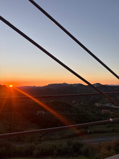 An outdoor scene depicts a sunset over the Unincorporated Santa Monica Mountains, United States of America. The sun, a distinct orange orb, is positioned on the horizon in the left third of the image, casting strong orange rays diagonally across the lower-left quadrant. The sky transitions from a bright orange at the horizon to a pale blue-grey at the top. Silhouetted mountains and hills covered in dark green vegetation dominate the midground, stretching across the width of the frame. Scattered among the hills, faint indications of roads and small structures, possibly buildings, are visible. Power lines traverse the middle distance horizontally. In the immediate foreground, three dark, parallel, linear objects, likely metal bars or wires, extend diagonally from top-left to bottom-right, partially obscuring the landscape. Two additional, similar linear objects are visible horizontally in the lower half of the frame. No individuals are present in the image.