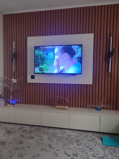 The image shows a modern living room interior with a television mounted on a wood-paneled wall. The wall features vertical wooden slats in a warm, reddish-brown tone, with thin black lines between them. A large, flat-screen television is centered on a white rectangular panel on this wall, displaying an image of a woman with her hand over her mouth, looking surprised or thoughtful. Two slim, vertical light fixtures are mounted on the wall, one on either side of the television.

Below the television is a long, white media console with three evenly spaced doors. On top of the console, from left to right, are a white vase with some decorative elements, a gold tree-shaped decorative object with small crystals and a blue light emanating from its base, a small, ornate gold basket, and a blue vase with wispy white decorative elements.

The floor is covered with a light-colored, patterned carpet. In the lower right corner of the image, a bright turquoise mat is visible on the floor. The overall impression is a well-lit, contemporary living space. There is no specific information in the image that would indicate its location in the city of Chuy, Kyrgyzstan.