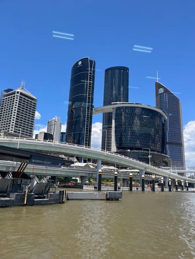 An urban waterside scene in Brisbane City, Australia, is captured during daytime under a bright blue sky with scattered white clouds. The image is composed of a wide river, a monorail track and station, and several modern skyscrapers.

On the right, a wide, murky brown-green river shows gentle ripples on its surface, indicating calm conditions. Along the riverbank, an elevated monorail track curves, leading to a station platform. The text "NORTH QUAY" is clearly visible on the side of the station, indicating its location. Below the monorail track and along the water's edge, docking structures and pylons are visible.

To the left of the monorail, several prominent skyscrapers dominate the skyline. These are mostly dark-colored and glass-clad, reflecting the bright sky. One distinctive building features the text "The Star" and a stylized starburst logo. Another building nearby clearly displays a "W Hotel" logo. Further buildings, varying in height and design, extend into the background. The clear sky in the upper left of the image shows some faint linear reflections or atmospheric phenomena. No specific human activity is depicted, but the presence of urban infrastructure suggests a bustling city environment.