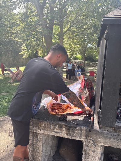 In Campezo/Kanpezu, Spain, a man with tattoos on his arm and leg is preparing to grill meat. He is wearing a black t-shirt and black shorts and is standing next to a large, dark grill. In front of him, on a concrete surface, is a pile of raw meat, mostly sausages, wrapped in a white paper bag with red and orange accents. He is using a tool to adjust something on the grill. In the background, other people are visible, some relaxing on hammocks and chairs, and others sitting at picnic tables. The scene is set in a grassy, tree-filled area, suggesting an outdoor recreational setting. The lighting indicates it is daytime, with sunlight filtering through the trees.