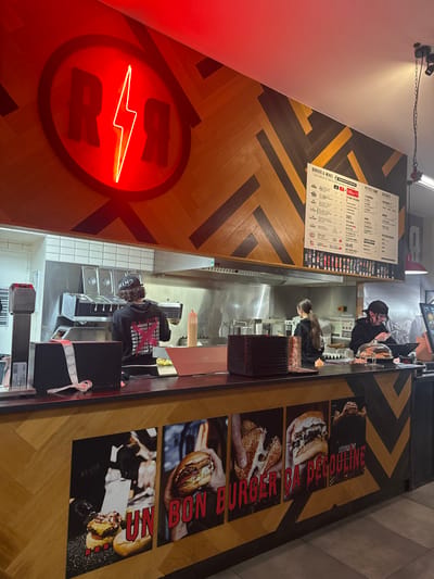 A brightly lit restaurant counter area. A neon sign with the letters "R R" and a lightning bolt is visible on the wall. The counter area is staffed by several employees, one wearing a baseball cap. The menu is visible on the wall. The floor is decorated with photos of burgers, each with a French caption.