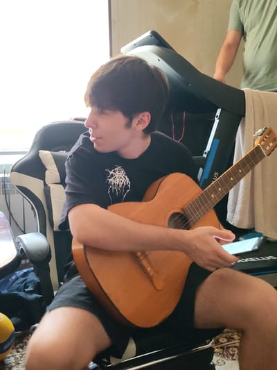 This media file features a young man sitting in a black and white gaming chair, holding an acoustic guitar. He is wearing a black t-shirt with a white graphic design and black shorts. His gaze is directed to his left, away from the camera. In his right hand, he is holding a smartphone, its screen illuminated. The guitar is made of light-colored wood and rests on his lap.
Behind the man, a treadmill is partially visible. A person in a green t-shirt is standing behind the treadmill, their arm resting on it. A white towel is draped over the treadmill.
The scene appears to be indoors, with natural light streaming in from a window on the left side of the frame. The background suggests a domestic setting. The overall mood is relaxed and casual. There is no clear indication of the time of day or weather.
Visible text includes the word "AKELLUS" on the treadmill. The provided location context is Yalangach, Uzbekistan.