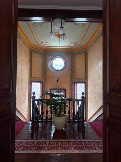 The image depicts the interior of a grand staircase landing within a historic building in La Orotava, Spain. The perspective is from an open doorway, looking into a brightly lit, opulent space. The landing features highly polished dark wood floors, partially covered by a deep red carpet with an intricate pattern. To the left and right, dark wooden banisters with turned spindles ascend the stairs, which are also carpeted in red.

The walls are adorned with textured panels in shades of peach and cream, framed by ornate gold moldings. Above, a decorative ceiling is painted with classical motifs in muted tones of green and gold. A prominent feature in the center of the landing is a large, potted green plant with broad leaves, placed in a textured white ceramic pot.

Dominating the upper portion of the scene is a circular window with a grid pattern, allowing natural light to filter into the space. Suspended from the ceiling by chains is a vintage light fixture with three spherical glass globes. The overall impression is one of refined elegance and historical preservation.