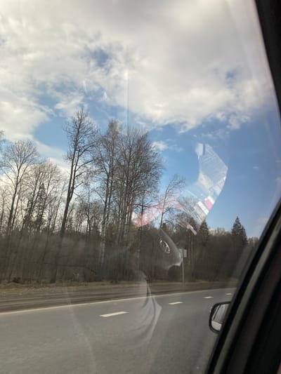 An outdoor scene is captured from inside a moving vehicle, looking out a side window. The image is oriented with the road appearing vertically on the right, and the sky on the left, suggesting a 90-degree clockwise rotation from a typical viewing perspective.

The foreground is dominated by reflections on the car window. Visible reflections include parts of the vehicle's interior, such as a dashboard or console, a side mirror, and a faint, blurry reflection of a person's arm or torso. A distinct reflection of a circular car emblem, possibly from a steering wheel, is also visible.

Beyond the reflections, the scene outside depicts a paved road with white dashed lane markings on the right. To the left of the road, a dense line of trees with bare branches extends along the roadside, primarily deciduous trees, suggesting late autumn, winter, or early spring. Further in the distance, a few evergreen coniferous trees are also visible among the bare ones. The sky is partially cloudy with patches of clear blue, indicating a bright, sunny day. Sun glare or lens flare is present in some areas, consistent with bright daylight conditions.

The activity depicted is travel by car along a road in what appears to be a rural or semi-rural setting. There is no discernible text visible in the image, and no specific emotions are conveyed. The location context for this scene is Bal'zyashur, Russia.