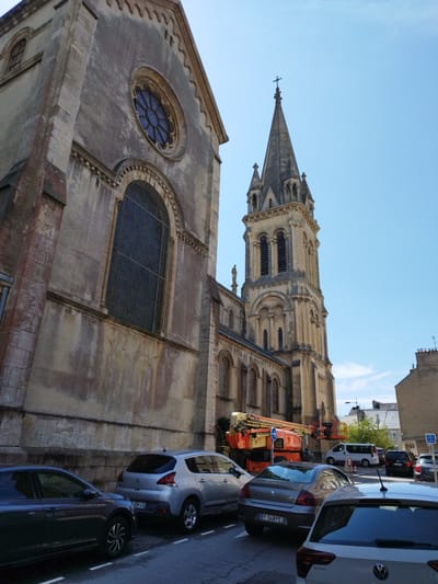 This image captures a street scene in Cherbourg-en-Cotentin, France, with a large stone church dominating the left side of the frame. The church features a prominent rose window and a tall, ornate bell tower topped with a cross, indicating a Gothic architectural style. The sky is a clear blue, suggesting a sunny day.
In the foreground, several cars are parked along the street. These include a dark-colored sedan, a silver hatchback, and the rear of a silver hatchback. Further down the street, other vehicles are visible, including a white van and a black car.
An orange aerial work platform, often used for construction or maintenance, is positioned on the street near the church, suggesting some ongoing work or repair. A blue directional sign with a white arrow is attached to the platform.
The architecture of the church, with its weathered stone and intricate details, along with the parked cars and the presence of the aerial platform, creates a sense of daily life in this French town, possibly during daytime hours. There is no visible text in the image other than the license plates on the cars. The license plates appear to be French, with formats like "459 LS" and "BY 548 FE".