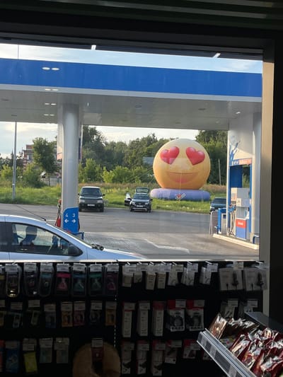 The image depicts a gas station with a large inflatable smiley face adorned with heart-shaped eyes situated in the background. Several cars are parked at the pumps, and a white car is visible in the foreground, partially obscuring a display of products. The gas station appears to be branded with "G-Drive 95." The overall scene suggests a commercial area, possibly in the city of Tri-Izby, Russia, given the context. The lighting indicates it is daytime, with sunlight casting shadows. The area surrounding the gas station features green trees and foliage.