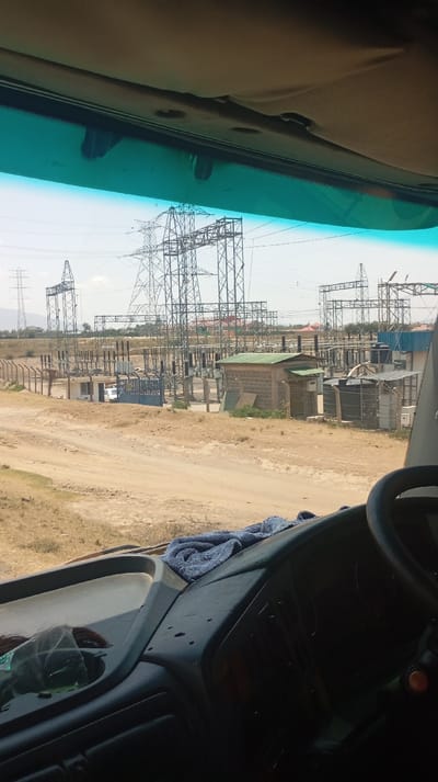 The image presents a daytime view from inside a vehicle, likely a truck or bus, looking forward through a tinted windshield. The lower portion of the frame shows the vehicle's dashboard, dark grey in color, with a portion of a steering wheel visible on the lower right. A crumpled blue towel rests on the dashboard towards the center-right.
Beyond the vehicle, a large electrical substation or power plant is visible under a clear, bright sky, consistent with the Hells Gate ward in Kenya. Numerous tall, grey metal lattice transmission towers are distributed throughout the scene, supporting overhead power lines. Various electrical equipment, including insulators and transformers, are arranged on platforms within the substation's fenced perimeter. Several small, single-story buildings are present within the complex; one has a green corrugated metal roof and appears to be constructed of cinder blocks, while another is white with a flat roof, possibly a guardhouse near a blue gate.
The ground surrounding the substation is predominantly dry, light brown earth, indicating an arid or semi-arid environment, with sparse, dry vegetation. Dirt roads and tracks crisscross the foreground and lead into the substation area. In the far distance, faint outlines of hills or mountains are visible under the horizon. No individuals or active movements are discernible in the scene. The overall impression is an industrial power infrastructure facility situated in a rural or developing area.
