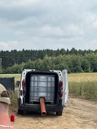 In Opatów, Poland, a white van is parked on a dirt road, its rear doors open. Inside the van, a large, grey IBC tote is visible, with a reddish-orange pipe extending from its bottom. The tote is positioned on a pallet. A person wearing a cap and a necklace is partially visible in the foreground, with a red object on the ground nearby. The van is situated in a rural setting, with a field of dry grass or crops behind it and a dense forest in the distance. The sky is overcast.