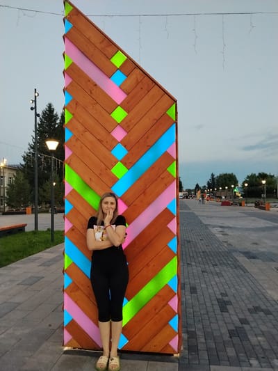 A blonde woman stands in front of a wooden structure adorned with colorful geometric shapes in Mikhailovka, Russia. The structure is located in a public space, possibly a park or a pedestrian street, with a paved walkway and streetlights visible in the background. The woman is wearing a black t-shirt and black leggings, with her arms crossed and her hand resting on her chin, suggesting a thoughtful or relaxed pose. The overall atmosphere of the image is calm, with the dusk sky indicating the time of day.
