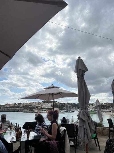 The scene depicts an outdoor dining or social area on a terrace overlooking the harbor of Porto Cristo, Spain. The sky is largely overcast with prominent cumulus clouds and visible patches of blue. A calm, blue-green body of water, likely a bay or inlet, occupies the midground, with a distant shoreline featuring light-colored buildings interspersed with green vegetation on a hillside. A marina filled with numerous moored boats and yachts is visible to the right, adjacent to a small breakwater with a red structure.
In the foreground and midground, several outdoor umbrellas are present. One light-gray umbrella is fully open, providing shade over a central table. Two other large, light-gray umbrellas are closed and folded upright in the right midground, and a corner of a fourth open umbrella is visible in the upper left foreground.
Four individuals are seated at a black rectangular table in the foreground. On the far left, an older male with gray hair, wearing sunglasses and a light-colored long-sleeved shirt, is seated with his left arm resting on the table. A hand and forearm, possibly belonging to another person, are visible to his right, gesturing over the table. Next, a female with dark, curly hair, wearing a striped long-sleeved shirt, looks down at a small object held in her hands. To her right, a female with reddish-brown hair tied in a ponytail, wearing glasses and a dark sleeveless top, looks towards her left. A purple/maroon jacket is draped over the back of her chair and partially covers her lap.
On the foreground table, multiple dark glass bottles, some empty and some partially filled, are visible alongside clear drinking glasses and small electronic devices. A partially visible table to the right holds additional glassware, a clear bottle, and a dark box-like object. The individuals are seated, engaging with items on the table and appearing to be in a social gathering.