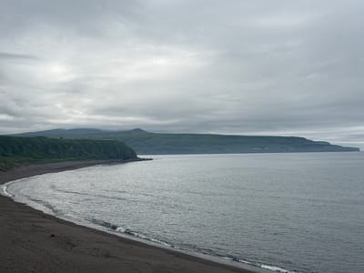 The image depicts a dark sand beach curving along the coast of a body of water. Gentle waves lap at the shore. Beyond the beach, a steep, green, treed cliff face rises. In the distance, a low-lying island or peninsula, also covered in greenery, stretches across the horizon. The sky is overcast with heavy, grey clouds. A small, light-colored structure is visible on the grassy area atop the cliff in the mid-left of the frame. The scene suggests a remote, coastal environment under a cloudy sky, likely on Sakhalin Island or the Kuril Islands, Russia, given the mention of Kuril'sk.
