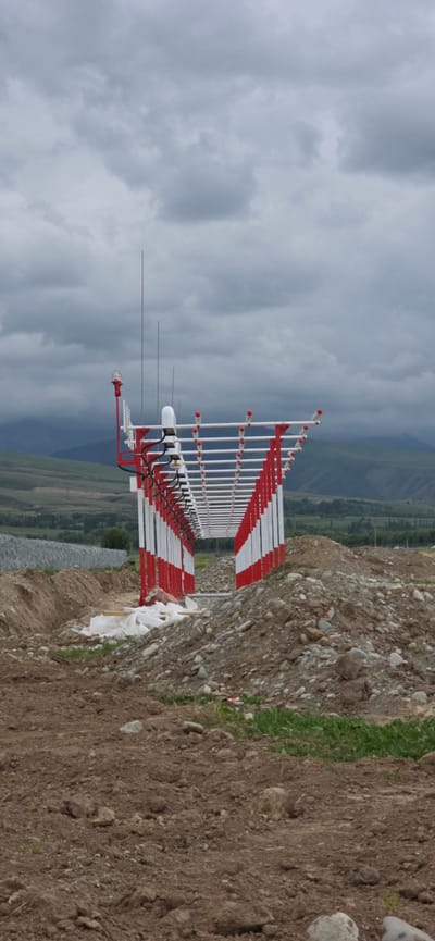 The image depicts an outdoor scene with a large, grid-like structure made of white and red metal poles, likely an antenna array or part of an airport navigation system, dominating the center of the frame. This structure is elevated and extends horizontally. To the left of the structure, there is a red and white signal light attached to a pole, along with several vertical antennas. The foreground is rough terrain with disturbed earth, piles of rocks and soil, and some white bags, suggesting construction or excavation. In the background, there are rolling hills and mountains under a cloudy sky. The location is identified as Maman, Kyrgyzstan. There are no people or animals visible in the image. The overall impression is one of an infrastructure project in a rural or semi-rural area.