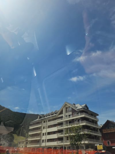 The image is a photo taken from a vehicle, likely a car, looking out at a bright blue sky with wispy clouds. There are reflections on the windshield, showing parts of the vehicle's interior, including a person's shoulder and head on the left side of the frame, and the top of what appears to be a dashboard or car frame in the upper portion of the image.
In the mid-ground, a multi-story apartment building with balconies and a pitched roof is visible. The building is light-colored with dark window frames. To the left of this building, a lush green mountain slope covered in trees rises sharply. Further to the right, a smaller, dark red building with wooden balconies is partially visible.
In the foreground, an orange construction fence is present, with a hint of a person in blue clothing to the far left. A yellow sign is also visible near the base of the apartment building. The overall scene suggests a daytime view of a town nestled against a mountainous backdrop, likely in the Italian Alps given the mention of Aprica, Italy.