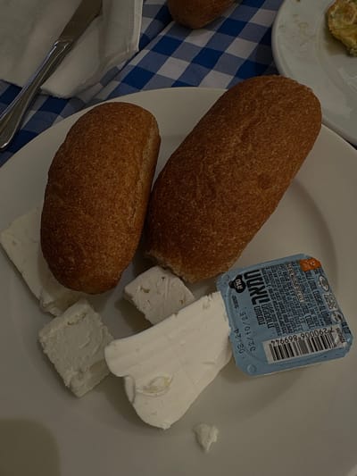 The image shows a plate with two rolls of bread, a small container of cream cheese, and a small piece of white cheese on a white plate. The plate is sitting on a blue and white checkered table cloth. The location is Ein Bokek, Israel at the coordinates 31.203 latitude and 35.361 longitude.
