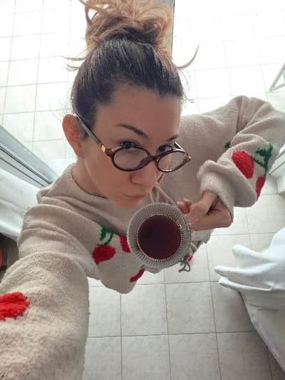 A female individual with dark brown hair styled in a messy bun and wearing brown, oval-framed eyeglasses with gold accents is captured from a high, overhead angle. She wears a cream-colored textured sweater featuring red cherry motifs with green stems on the sleeves and torso, along with a thin gold-colored necklace. Her right hand extends forward, holding a clear glass cup with a silver-colored, ribbed rim, containing a dark reddish-brown liquid. Her gaze is directed towards the camera. The floor beneath her consists of light-colored square tiles. The background is predominantly bright white, suggesting strong ambient light or an outdoor view, with a dark horizontal line visible on the upper left, possibly a window or door frame. A partially obscured white, soft object is visible in the lower right background. The scene is located in the Municipal Unit of Corinth, Greece.