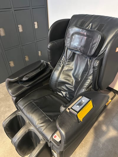 A black leather massage chair is positioned in front of a row of gray lockers. The chair has a headrest, armrests, and a footrest area. A yellow instruction label is attached to the right armrest. The lockers have rectangular openings for handles. The floor is tiled with a light-colored material. The lighting is ambient.