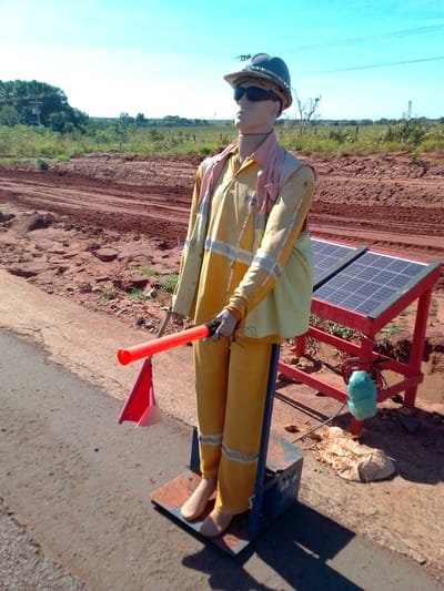 The image shows a mannequin dressed as a construction worker, standing on a roadside. The mannequin is wearing a yellow uniform with reflective stripes, a hard hat, and sunglasses. It is holding a red traffic baton. Behind the mannequin, there is a red wooden stand with two solar panels. The scene appears to be a rural area with a dirt road and vegetation in the background. The sky is clear and blue. There is no indication of the specific location within São Gabriel Do Oeste, Brazil.