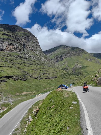 This image depicts a scenic mountain road scene in Koksar, India. A person on a motorcycle, loaded with luggage, travels along the asphalt road. The road curves to the left, with a steep, grassy slope on the right and a lower, greener valley on the left. Mountains covered in green vegetation and rock formations dominate the background. A blue and grey tent is pitched on the left side of the road, near a signpost. The sky is a vibrant blue with scattered white clouds.