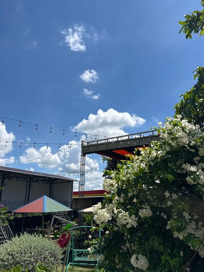 This image depicts an outdoor recreational area under a bright blue sky with scattered white clouds, likely in Ia Hru, Vietnam. In the foreground, a lush green bush with white flowers dominates the right side of the frame. To the left, a light green shrub with small leaves is visible, bordering a colorful, multi-sided umbrella-shaped roof structure that appears to be a pavilion. Adjacent to the pavilion is a teal-colored swing set with a red seat.

In the mid-ground, a multi-story structure with a dark roof and white walls is partially visible on the left. Above this, a raised walkway or balcony, supported by metal beams and featuring a visible orange awning underneath, stretches across the frame. Strings of exposed light bulbs are suspended in the air between poles and structures, suggesting this is a place for evening gatherings. In the far background, a building with a red and yellow facade can be seen. The overall scene suggests a well-maintained outdoor cafe or park setting. There are no people visible in the image.