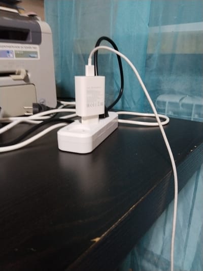 This media file shows a close-up view of a desk surface with various electronic devices and cables. In the foreground, a black desk is visible, with a white power strip plugged into the wall, which has a white phone charger plugged into it. A white USB cable and a black cable are connected to the charger, with the white cable extending out of frame. Behind the charger, a light-colored printer is partially visible. The background is a blurry teal curtain. There is no indication of the time of day or weather. The scene appears to be indoors, likely in a home or office setting. The visible text on the printer reads "USB 2.0" and what appears to be Russian text. The charger has "EAC" and "CE" markings.