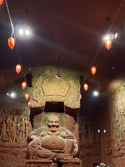 The image shows a stone Buddha sculpture in a dark environment, possibly inside a grotto or cave. The sculpture is ornately carved with detailed features, including the Buddha's face, robes, and surrounding patterns. Red lanterns are strung along wires, illuminating the scene. The sculpture is located in Shizhong, China. A person is visible on the right side of the frame, observing the statue.