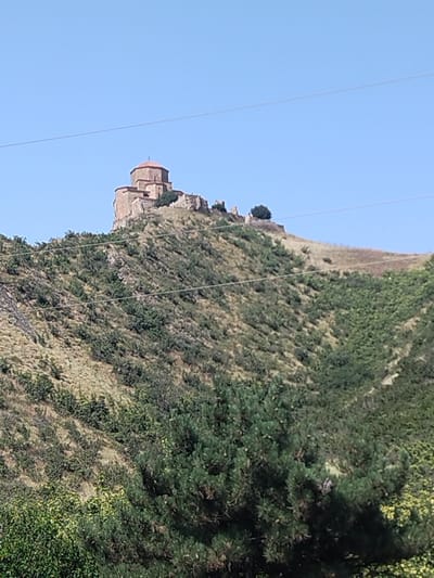 The image shows a historic monastery complex situated on a hilltop under a clear blue sky. The monastery features stone structures with a prominent, reddish-brown dome. The hillside is covered in a mix of dry grass and dense green shrubbery, with some trees visible in the foreground. The scene is characterized by its natural landscape and the ancient architecture of the monastery. The image captures a serene and picturesque view of the location.