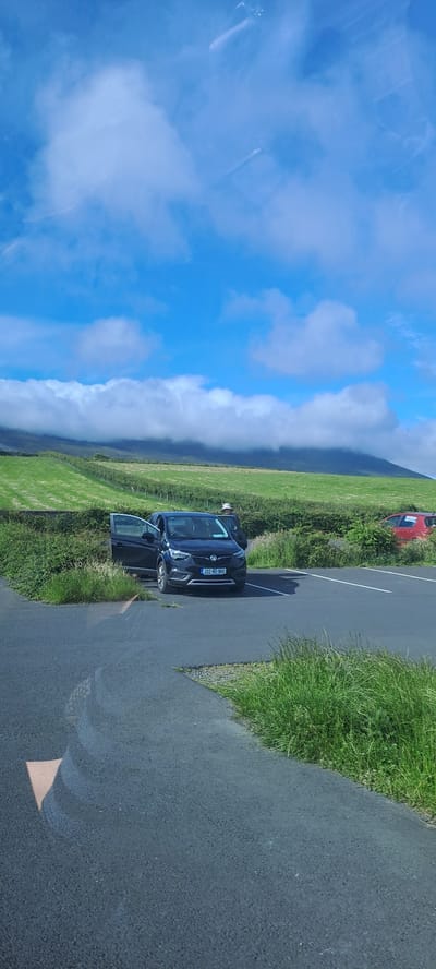 A black Vauxhall Crossland X SUV with its driver's side door open is parked in a parking lot. The car's license plate reads "202-HO-960". A person wearing a light-colored hat stands near the open driver's side door. Another red car is parked further down the lot, also with a door slightly ajar. The parking lot is paved with asphalt and bordered by tall green grass. In the background, there are terraced green fields leading up to a misty, cloud-covered hillside. The sky is bright blue with scattered white clouds. The location is Pier Road, Ireland.