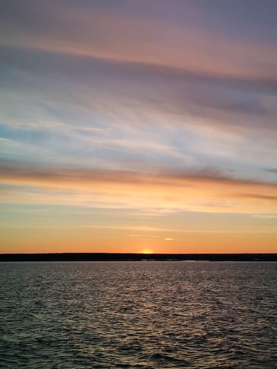 The image captures a sunset over water, likely a lake or sea. The sky is filled with bands of color, ranging from pale blue to orange and purple. At the horizon line, a dark stretch of land is visible, with the sun setting directly behind it. The water reflects the colors of the sky, creating a shimmering effect. The location is indicated as Vasil'evskoe, Russia, suggesting a natural landscape rather than a dense urban area. No people or other specific objects are visible in the frame.