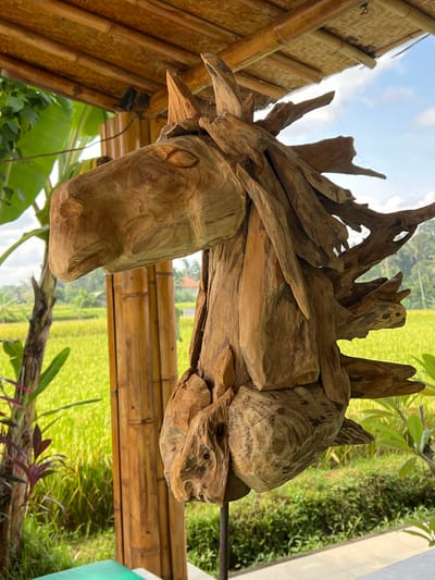 The image shows a wooden sculpture of a horse's head. The sculpture is made from pieces of wood, giving it a rough and textured appearance. It is situated outdoors, hanging from a bamboo structure with a rice field visible in the background. The location is Singakerta, Indonesia.
