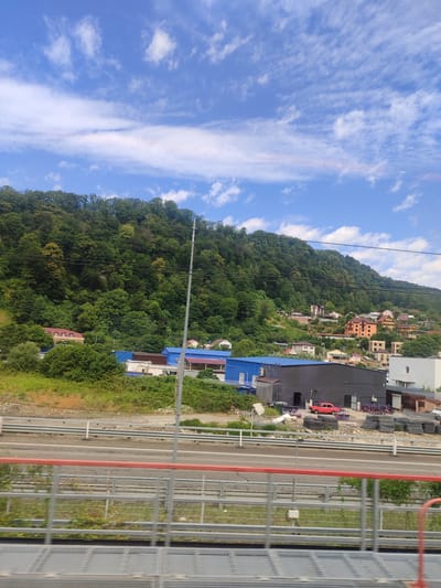 The image depicts a bright, sunny day with a partially cloudy blue sky above a verdant, tree-covered hillside. The hillside is dotted with houses of varying colors and architectural styles, nestled amongst the trees. In the foreground, a multi-lane road runs horizontally across the frame, separated by a barrier. Adjacent to the road are industrial-looking buildings, one with a blue roof and another dark gray structure. A red car is parked in front of the dark gray building. Beyond the road and buildings, a metal railing system is visible, suggesting the image was taken from an elevated position, possibly a bridge or overpass. The area appears to be a semi-urban or suburban environment bordering a natural landscape. The city of Kazachiy Brod, Russia, is characterized by its hilly terrain and proximity to forested areas, consistent with the scene depicted.