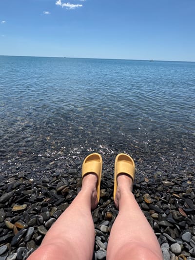 The image depicts a person's legs and feet, adorned in yellow slip-on sandals, standing on a dark, pebbly beach. The gentle waves of the sea lap at the shore, with the water appearing clear and shallow, revealing the submerged stones. The horizon is a distinct line separating the blue sky from the water, with a small, distant silhouette of what appears to be a boat or a person on a vessel. The sky is a clear, bright blue, suggesting a sunny day. The light glinting off the wet pebbles and the water indicates it is likely daytime, possibly mid-morning or afternoon given the brightness. The overall scene evokes a sense of relaxation and a casual summer day at the beach. The image orientation is vertical, suggesting it might have been taken while standing or in a portrait mode. There is no visible text in the image. The location context provided is Bol'shoi Utrish, Russia.